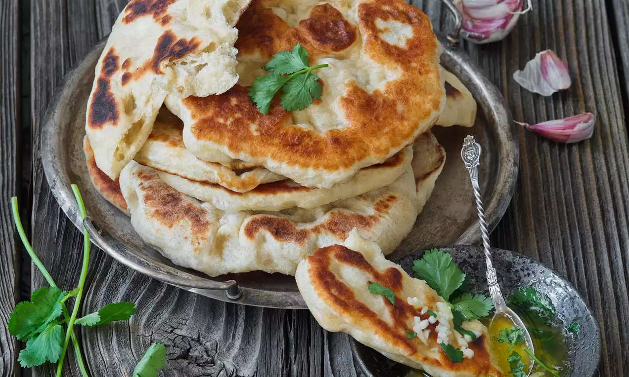 Zlatavé placky naan s lehkými hnědými opálenými skvrnami, měkká textura, vrstvené na kovovém talíři s čerstvým koriandrem a rozpuštěným máslem