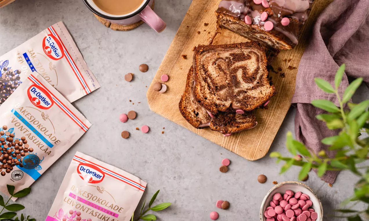 Marmurinis pyragas su šokolado raštais, padengtas blizgia glaisto pluta, pjaustytas riekėmis ant medinės lentelės, apsuptas spalvotų šokolado dražė ir rožinių dekoracijų, žalumos ir kavos puodeliu šone