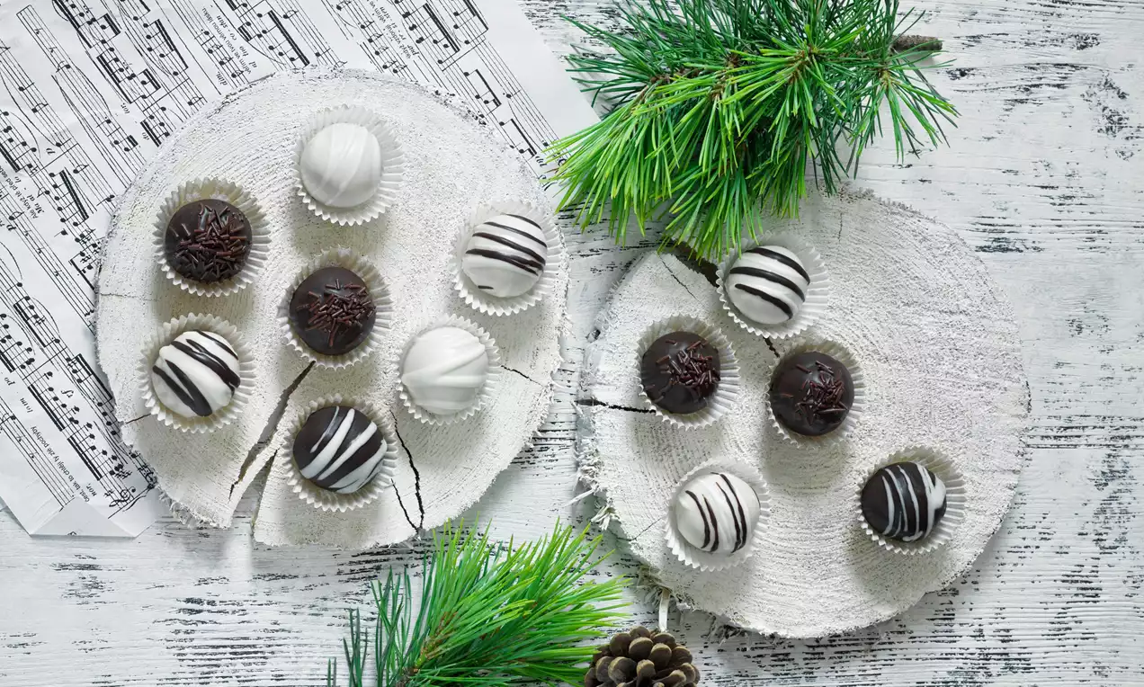 Tmavé a bílé marcipánové kuličky s texturami čokoládových proužků a hoblinek na bílých dřevěných podložkách vedle zelených větviček a šišky