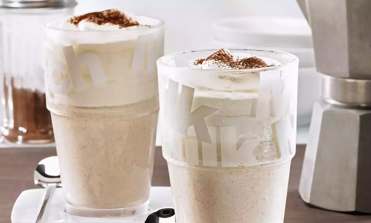 Deux verres glacés givrés remplis de crème latte beige, recouverts de mousse blanche et saupoudrés de cacao brun sur une table.