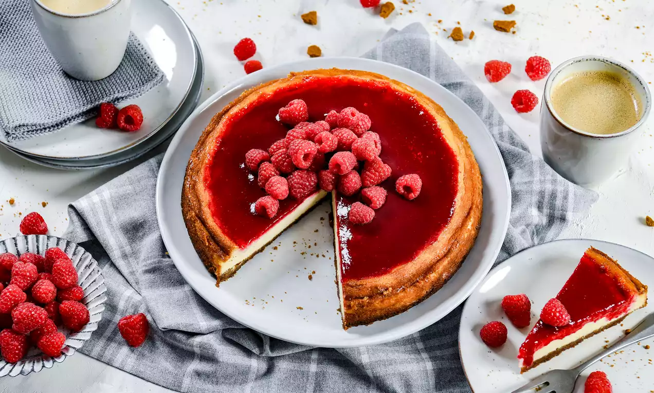 Krēmkrāsas siera kūka ar gludu aveņu glazūru, dekorēta svaigām avenēm centrā, uz balta šķīvja ar pelēku auduma akcentu