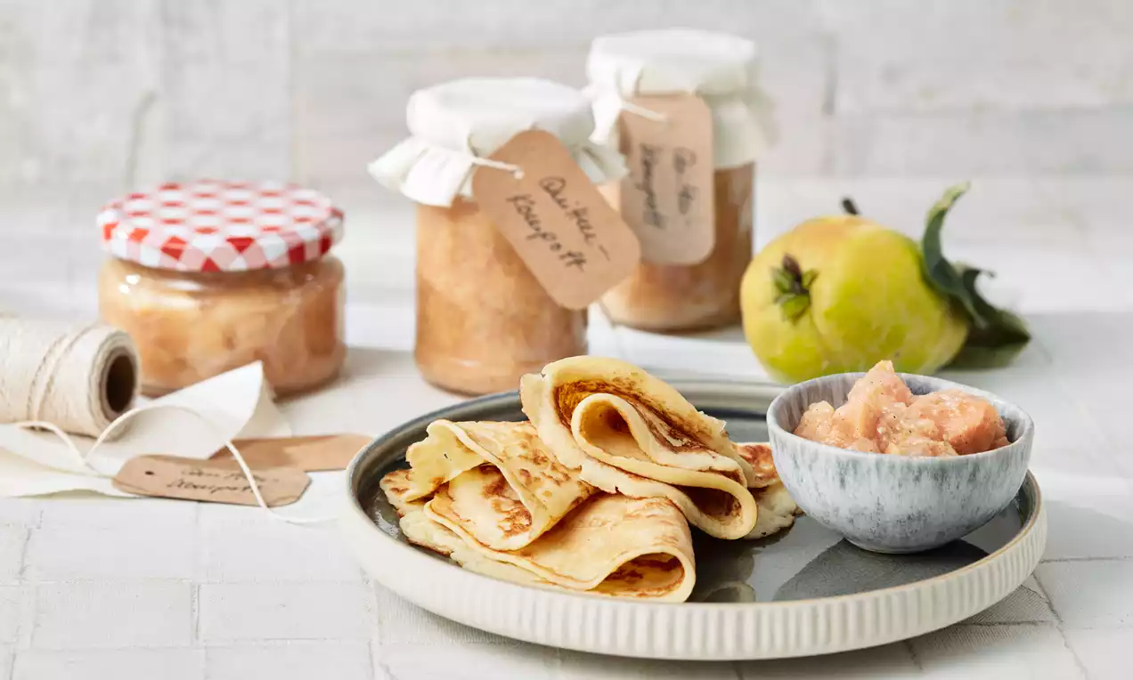 Gefaltete goldgelbe Pfannkuchen auf einem grauen Teller, daneben Quittenkompott in einer grauen Schale, umgeben von Gläsern und einer frischen Quitte