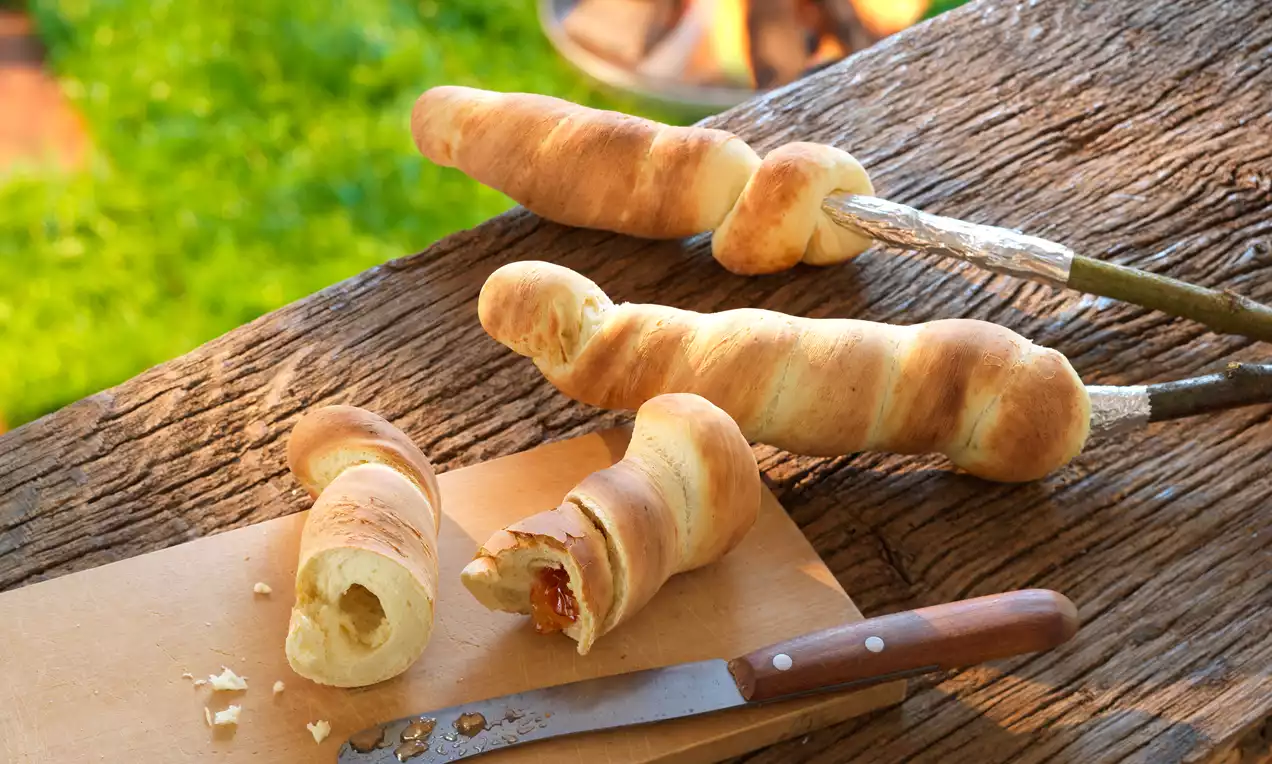 Zlatavé spirálovitě kroucené pečivo na špejli, jedno rozkrojené na dřevěném prkénku s nožem, v pozadí tráva a rozžhavený oheň