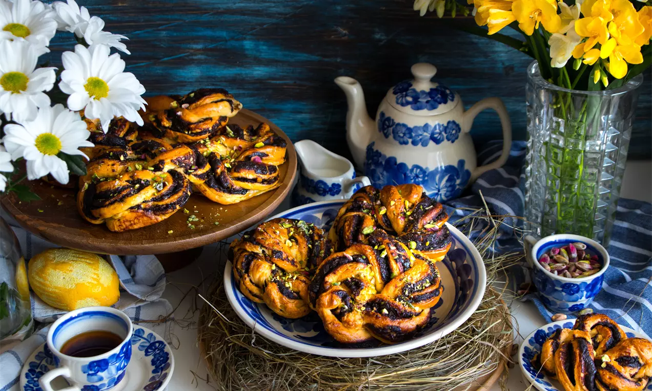 Златисто-кафяви плетени питки с лъскава повърхност и теменужени нишки, подредени в чинии върху сено до синьобелити порцеланови съдове