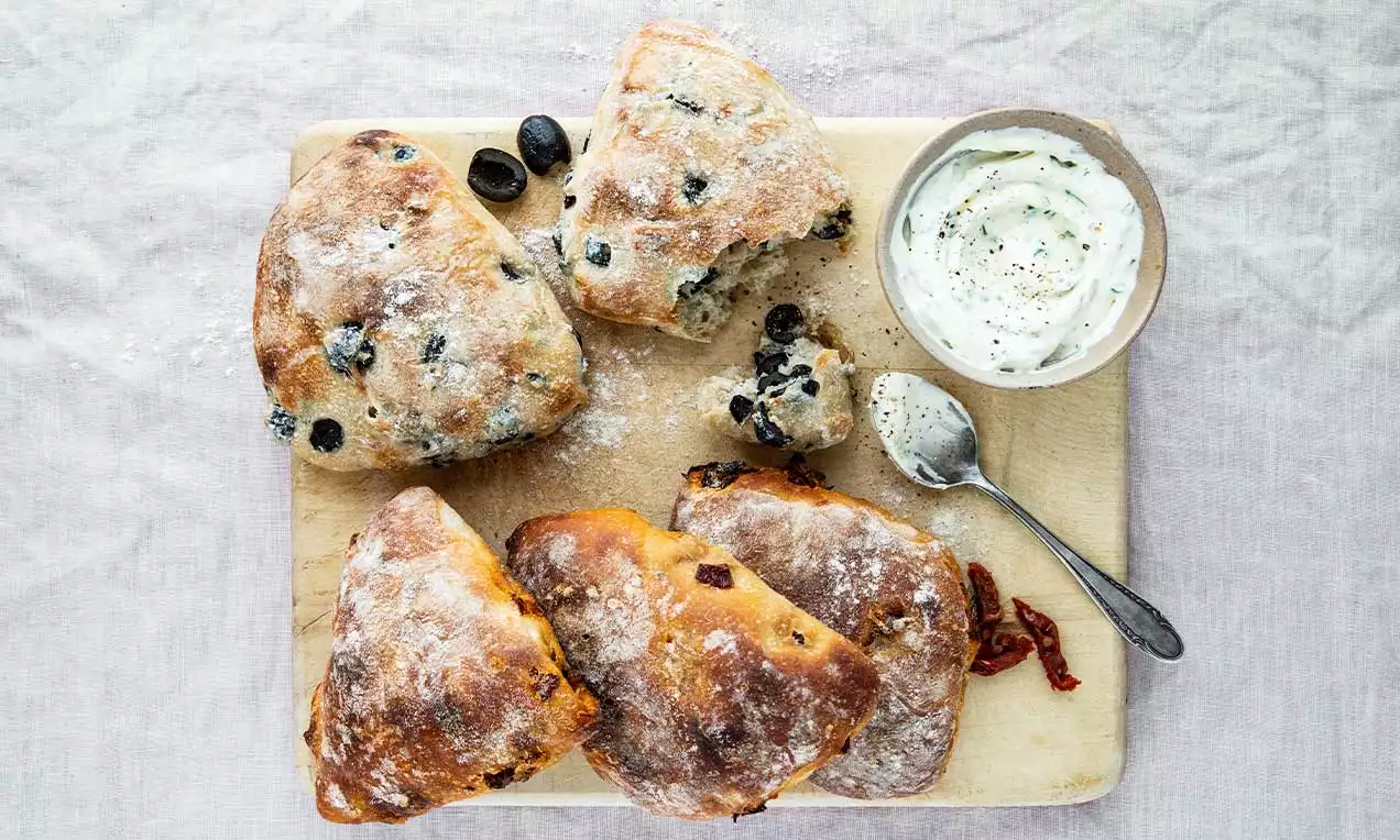 Oliven- und Tomaten-Brötli mit goldbrauner Kruste und Mehl bestäubt, auf Holzbrett angerichtet, daneben weißer Dip und getrocknete Tomaten