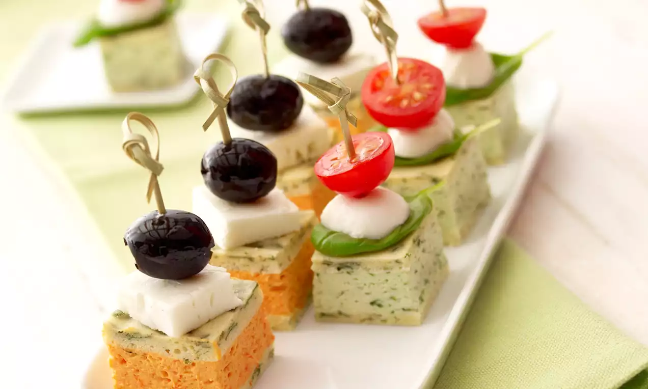 Barevné vrstvy kostek z bylinkového a paprikového krému, ozdobené cherry rajčaty, olivami a sýrem, napíchnuté na ozdobných špejlích
