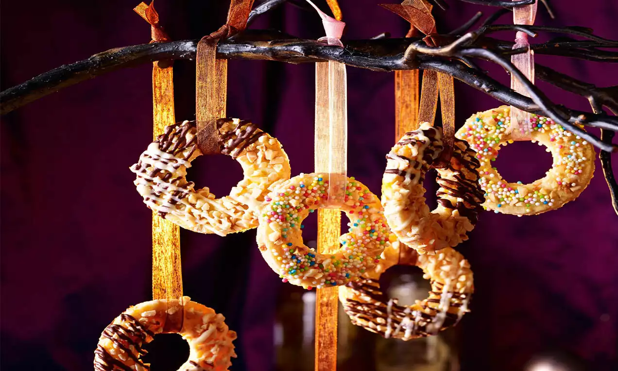 Kulaté křupavé kroužky zlatavé barvy, zdobené čokoládou, barevným posypem a zavěšené na stuhách na větvi tmavého stromu