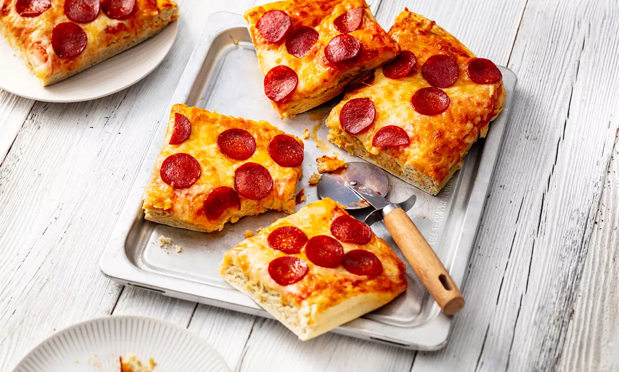 Американска пица с дебела правоъгълна кора, златисто препечена, покрита с бледожълта разтопена моцарела и кръгли резени червеникаво пеперони