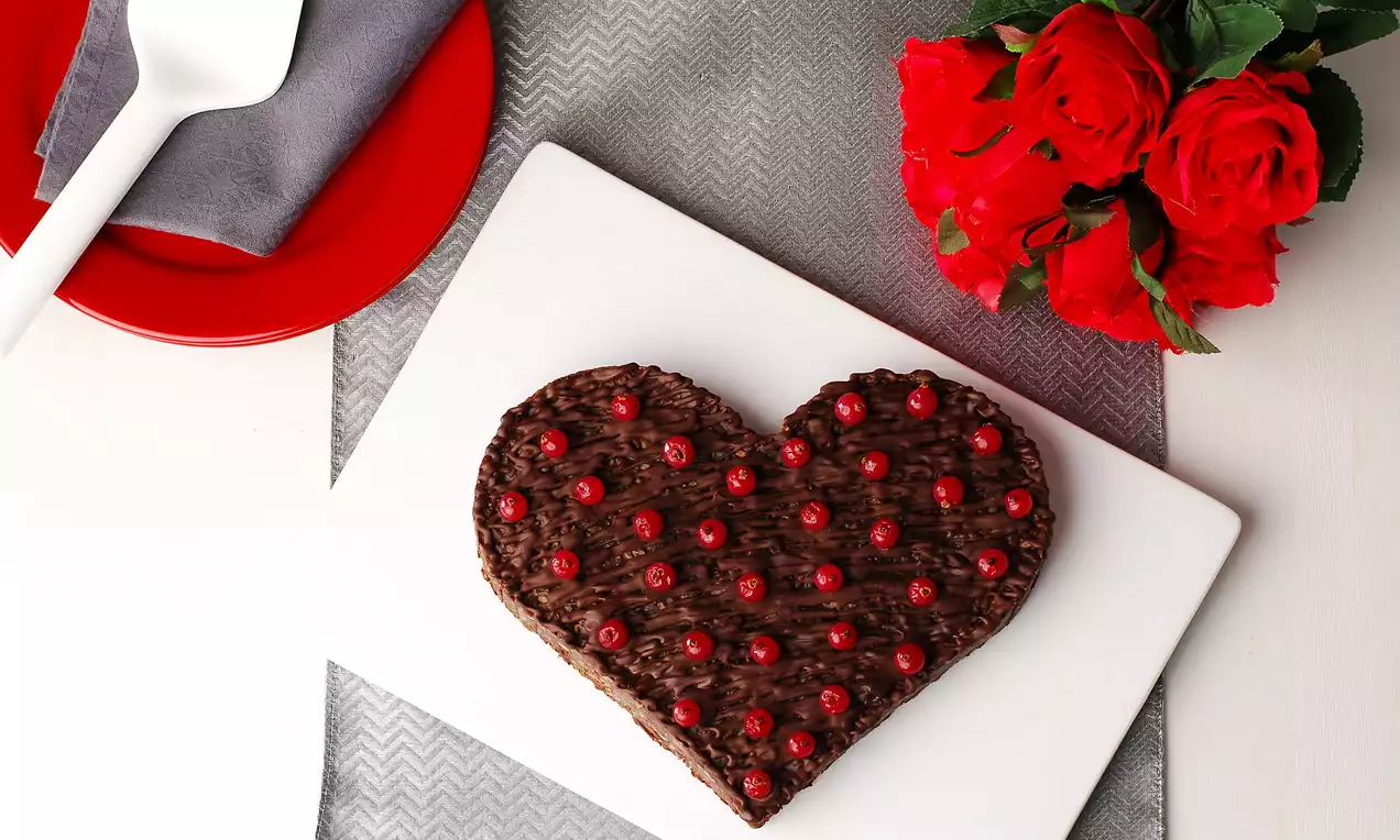 Kalp şeklinde çikolatalı tatlı, kırmızı meyvelerle süslenmiş, beyaz dikdörtgen bir tabakta, gri örtü ve kırmızı güllerle sunulmuş