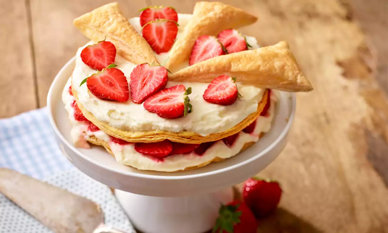 Torta iz zlato rjavega listnatega testa, premazana s puhasto belo kremo, obložena s svežimi jagodami in dekorirana s trikotniki testa na belem podstavku