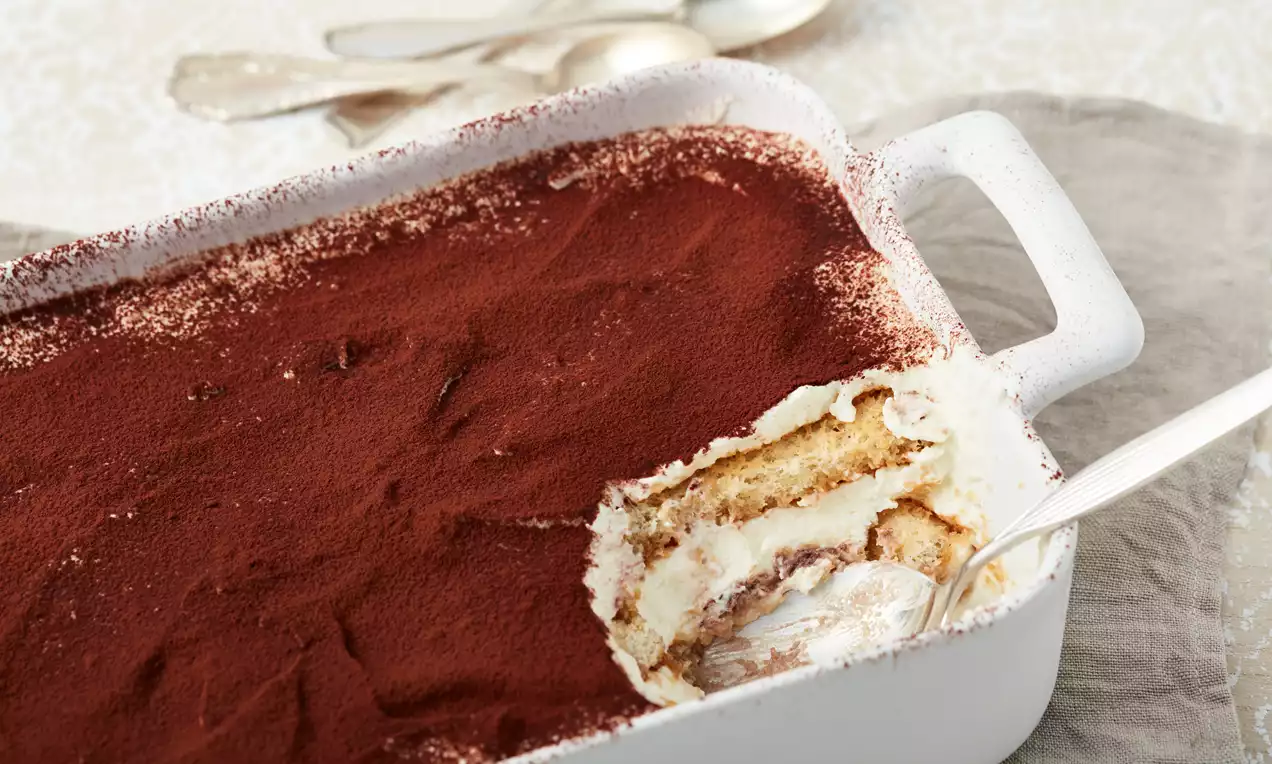 In einer weißen rechteckigen Form mit Löffel angerichtetes Tiramisu, mit dunkelbraunem Kakao bestäubt und locker geschichteter Creme und Biskuit