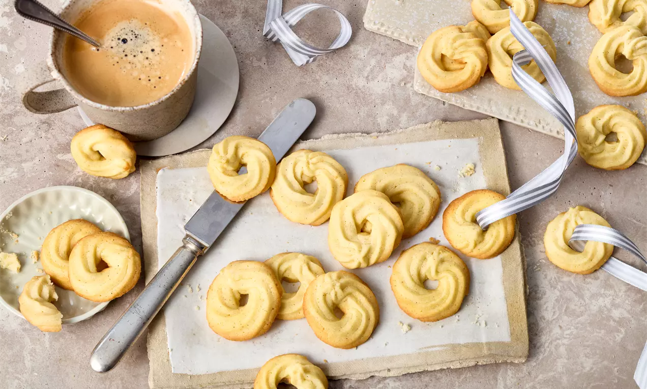 Goldgelbe Butter-Ringe mit leicht gebräunter Oberfläche auf Backpapier, daneben ein Messer, eine Tasse Kaffee und weiße Geschenkband-Dekoration