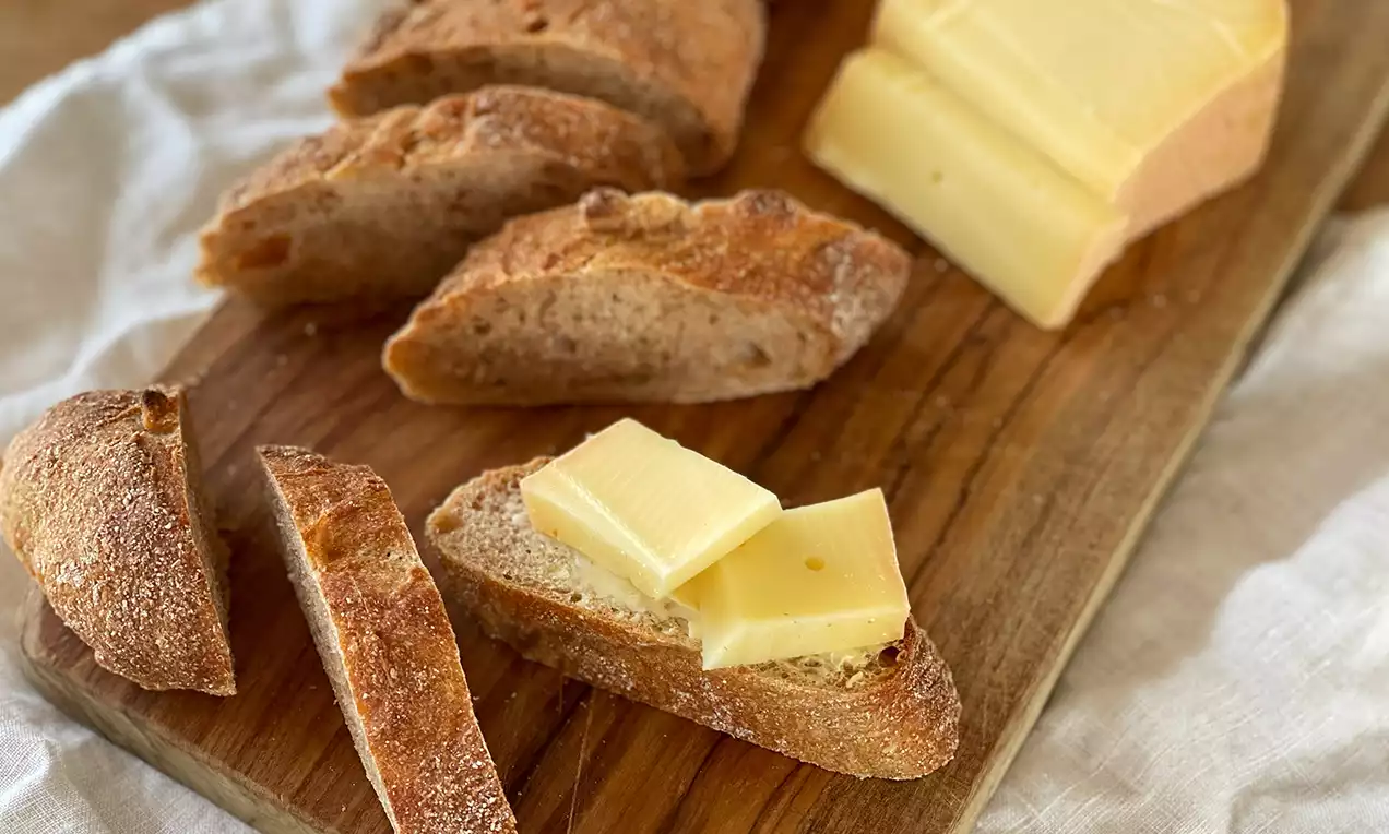 Dinkelbaguette mit goldbrauner Kruste in Scheiben geschnitten, auf Holzbrett mit zwei Käsestücken, eine Scheibe mit Butter belegt