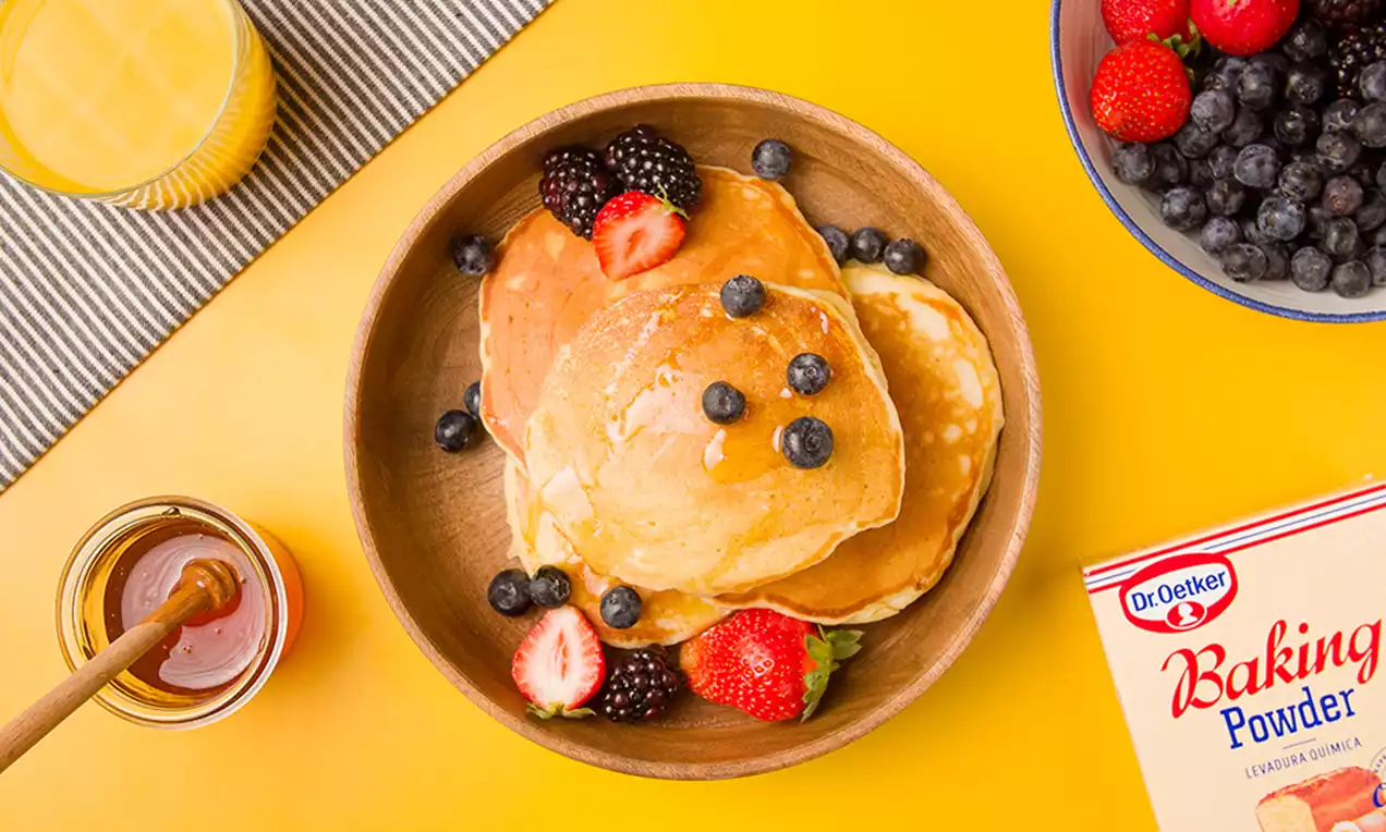 Tortitas doradas y esponjosas dispuestas en un cuenco de madera, decoradas con fresas, moras y arándanos sobre mesa amarilla
