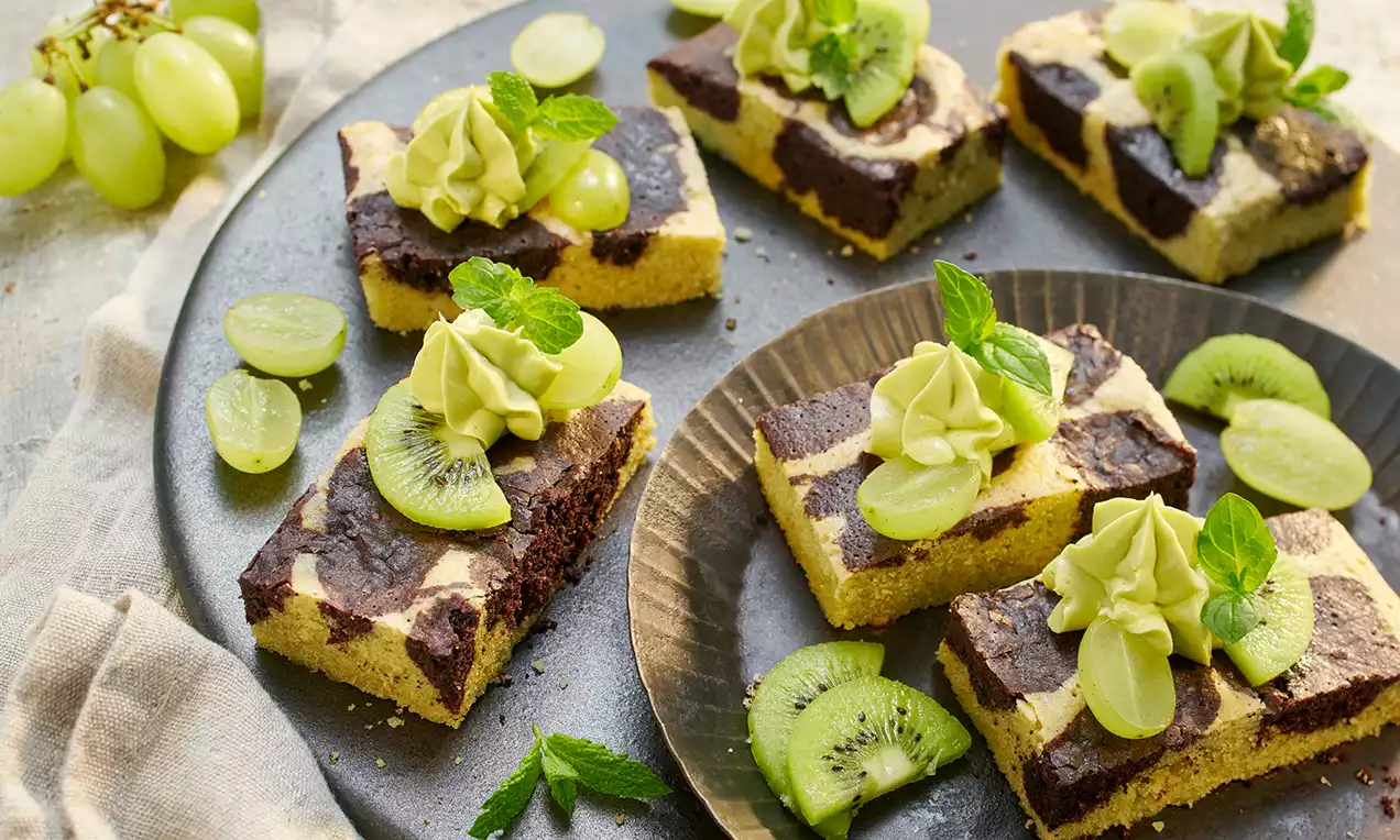 Vegane Marmor-Brownies mit dunkelbraunem Schokoladenmuster auf hellem Teig, dekoriert mit grünem Matcha-Topping, Kiwi und Weintrauben