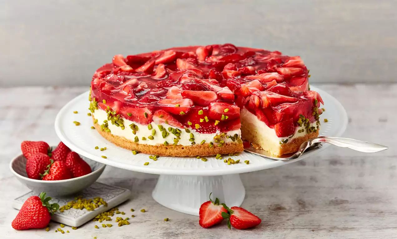 Runde Erdbeertorte mit glänzend rotem Belag aus frischen Erdbeerscheiben und Tortenguss, bestreut mit gehackten Pistazien auf weißer Tortenplatte