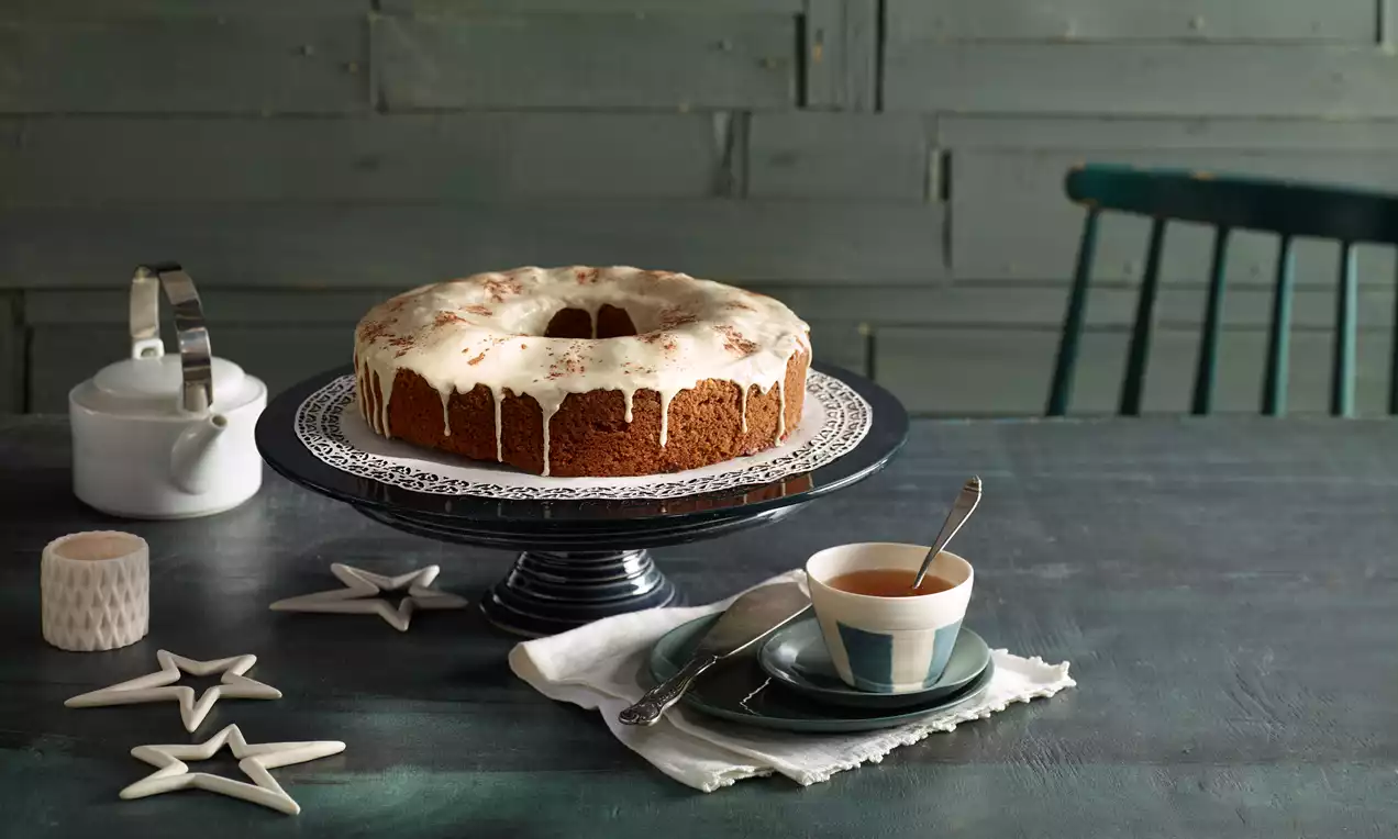 Kranzförmiger Gewürzkuchen mit glänzendem weißem Zuckerguss auf dunkler Tortenplatte, daneben Teetasse und Dekoration mit Sternen