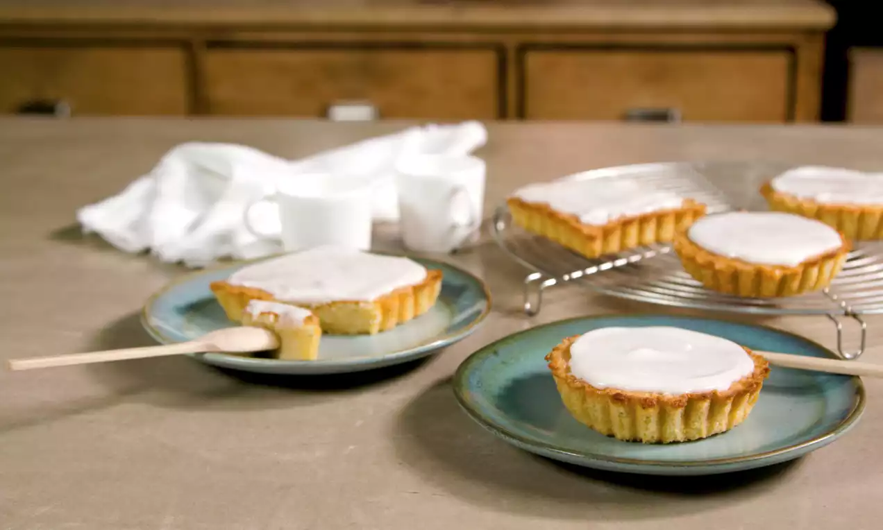 Frangipanetaartjes met goudgele korst en witte glazuurlaag op blauwe borden en een metalen rooster, geserveerd op een houten tafel