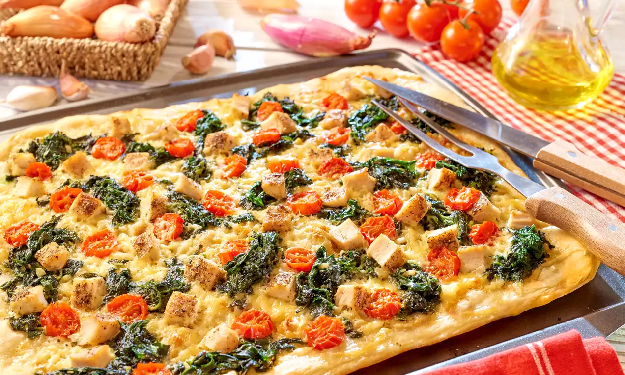 Prostokątna pizza na puszystym, złocistym cieście z kawałkami kurczaka, liśćmi szpinaku i plastrami pomidorów koktajlowych na blasze