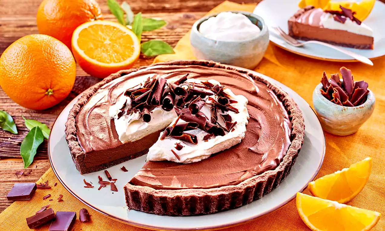 Tarta z ciemnoczekoladowym nadzieniem na kruchej brązowej podstawie, udekorowana bitą śmietaną, wiórkami czekolady i plasterkami pomarańczy