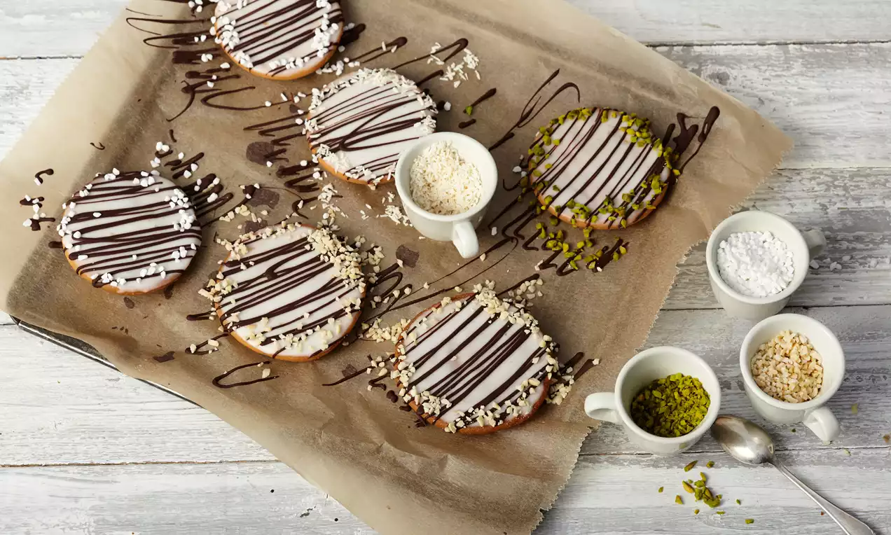 Runde Amerikaner mit weißer Glasur, dunklen Schokoladestreifen und gehackten Nüssen auf Backpapier, daneben Schälchen mit Zutaten