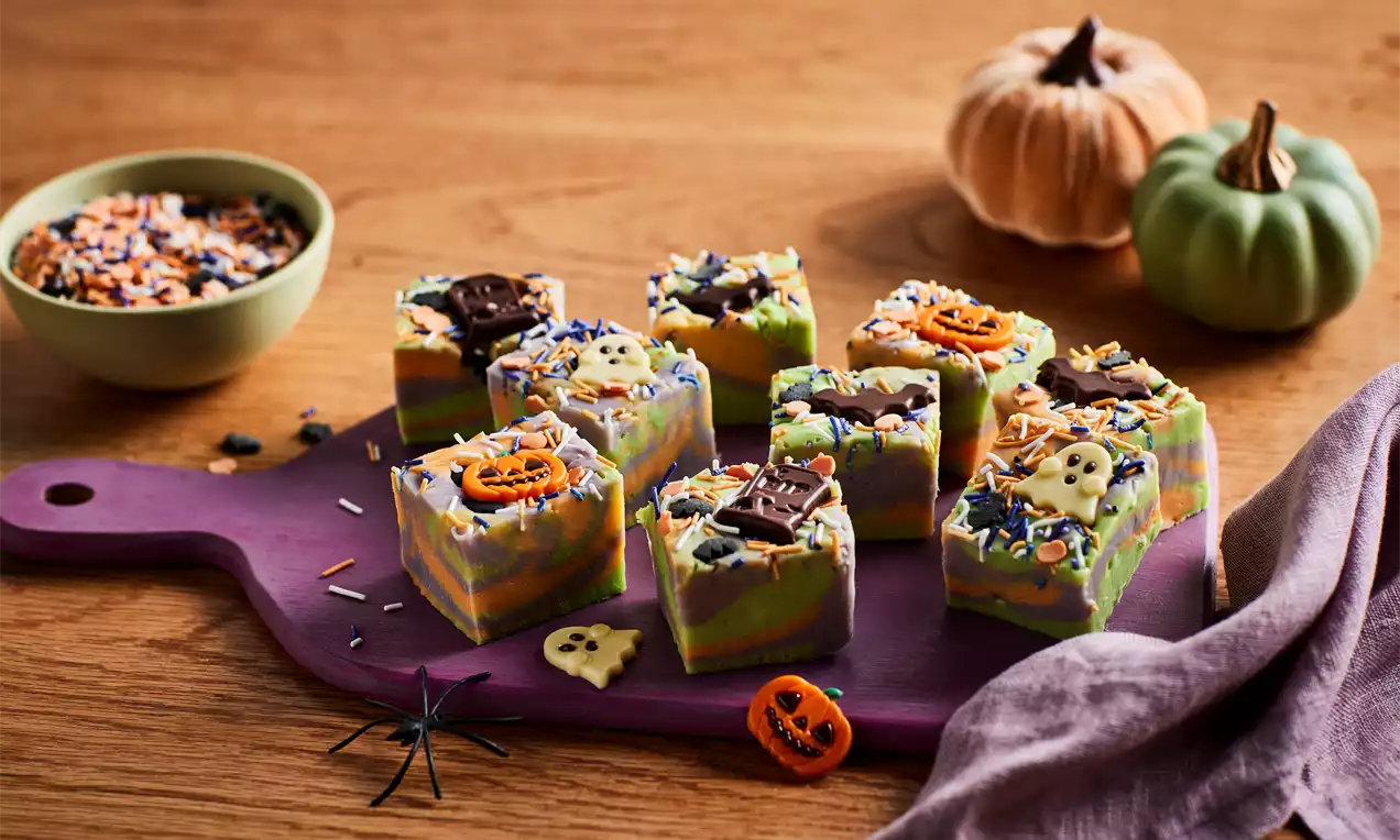 Colourful Halloween fudge squares with marbled orange, green and purple layers, sprinkled with candy pieces and chocolate shapes on a purple board