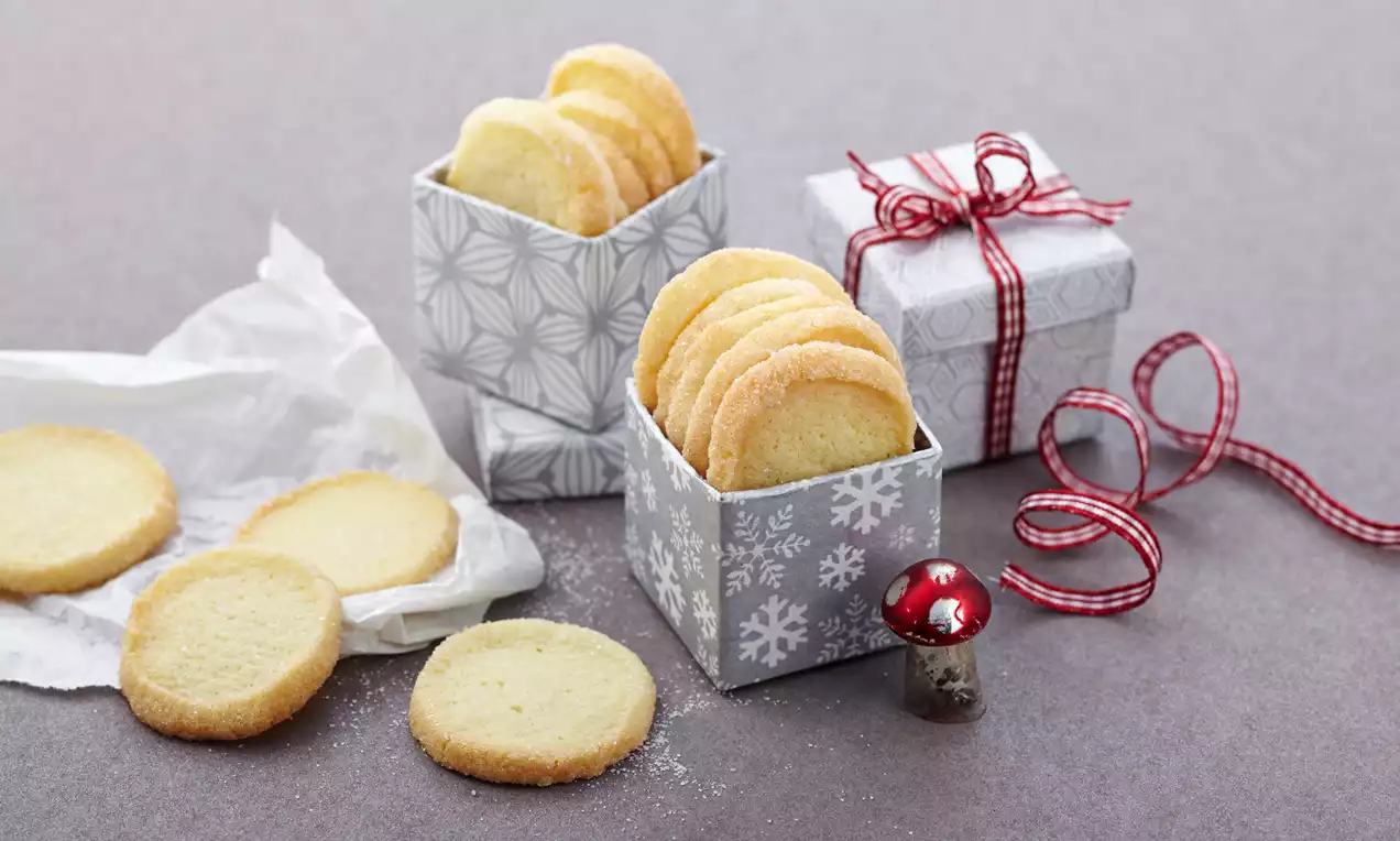 Heidesand-Plätzchen, goldbraun und mürbe, mit Puderzucker bestäubt und teils gestapelt auf weißem Teller auf rustikalem Holzbrett