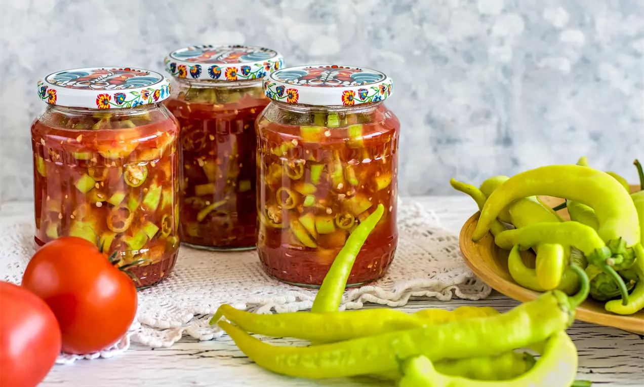 Чорбаджийски чушки с яркозелен цвят, нарязани и потопени в наситено червен доматен сос, поднесени в стъклени буркани на бяла покривка