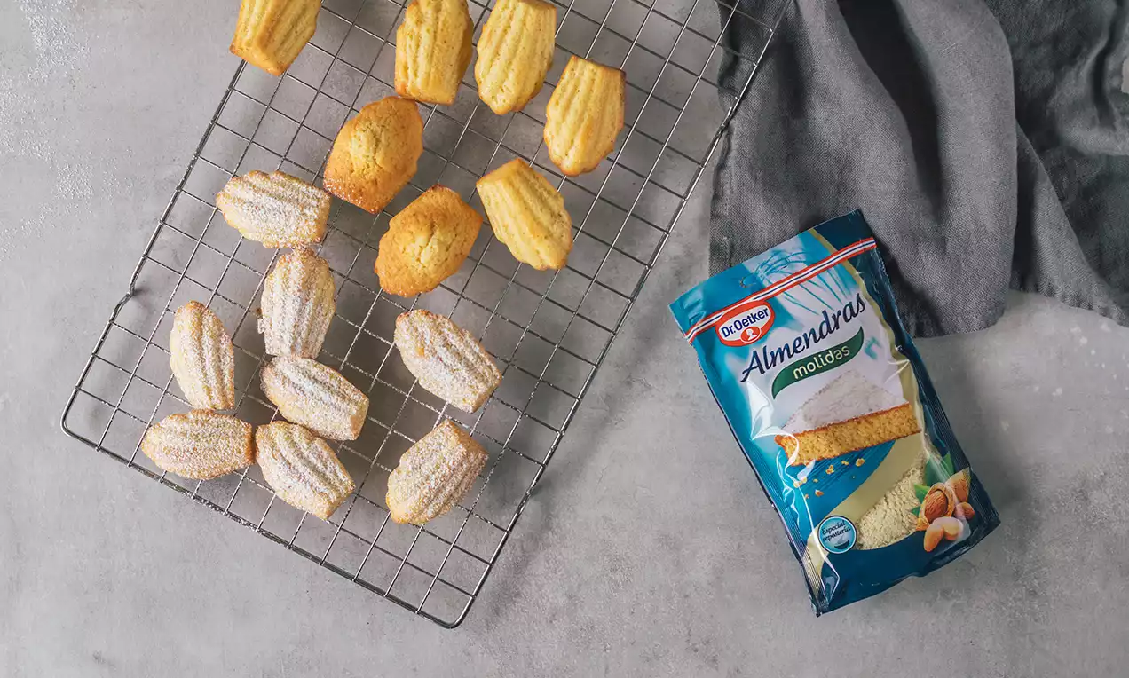 Magdalenas doradas con forma de conchas, textura rayada y esponjosa, espolvoreadas con azúcar glas sobre rejilla metálica junto a un paño gris