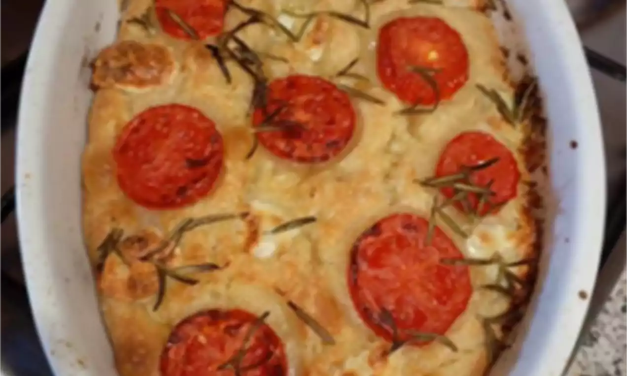 Zlatnosmeđa focaccia mekane, neravne teksture, ukrašena cijelim kriškama crvenih rajčica i grančicama svježeg ružmarina u bijelom ovalnom kalupu