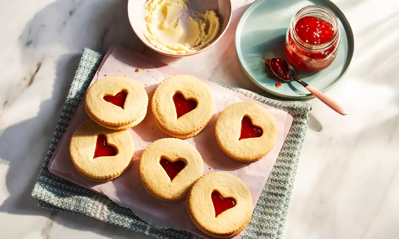 Golden vanilla biscuits sandwiched with red jam, heart-shaped cutouts on top, arranged in rows on pink parchment paper