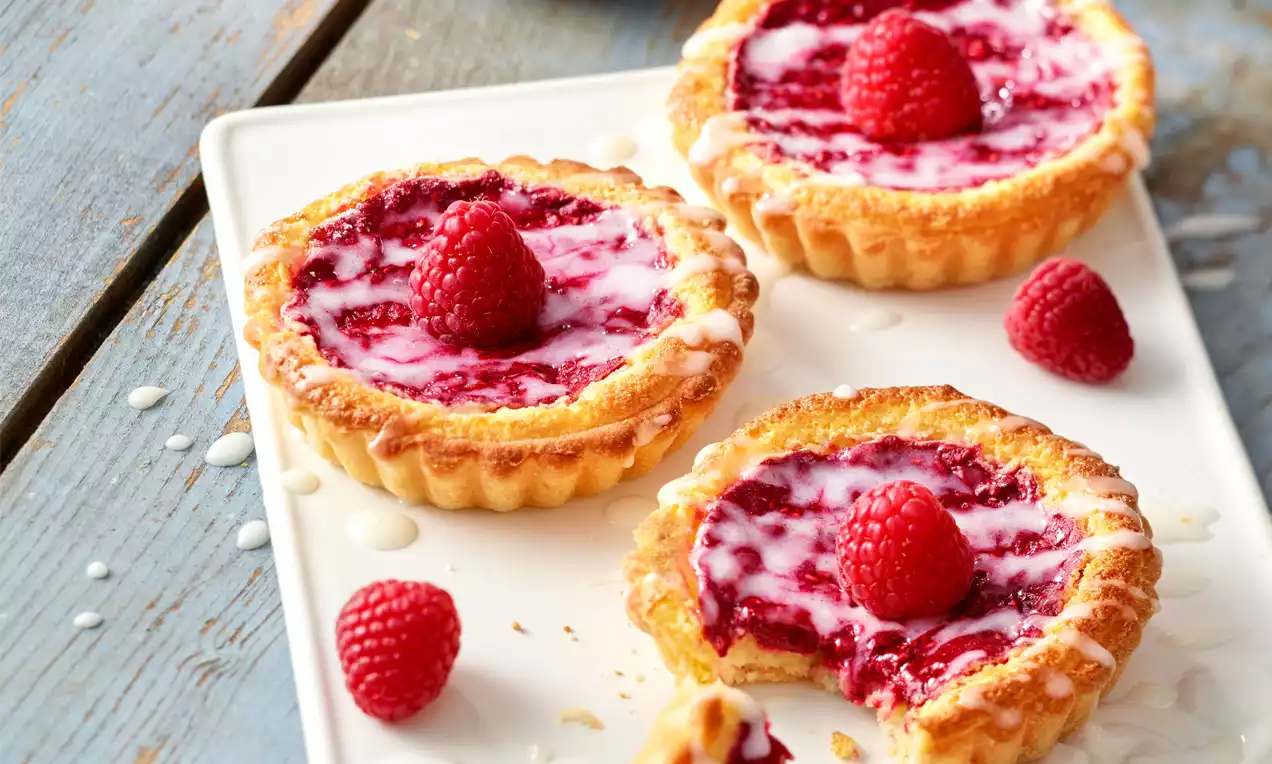 Kleine Cheesecake-Tartelettes mit goldbraunem Mürbeteig, himbeerrotem Fruchtspiegel, Zuckerguss und frischen Himbeeren auf weißer Platte