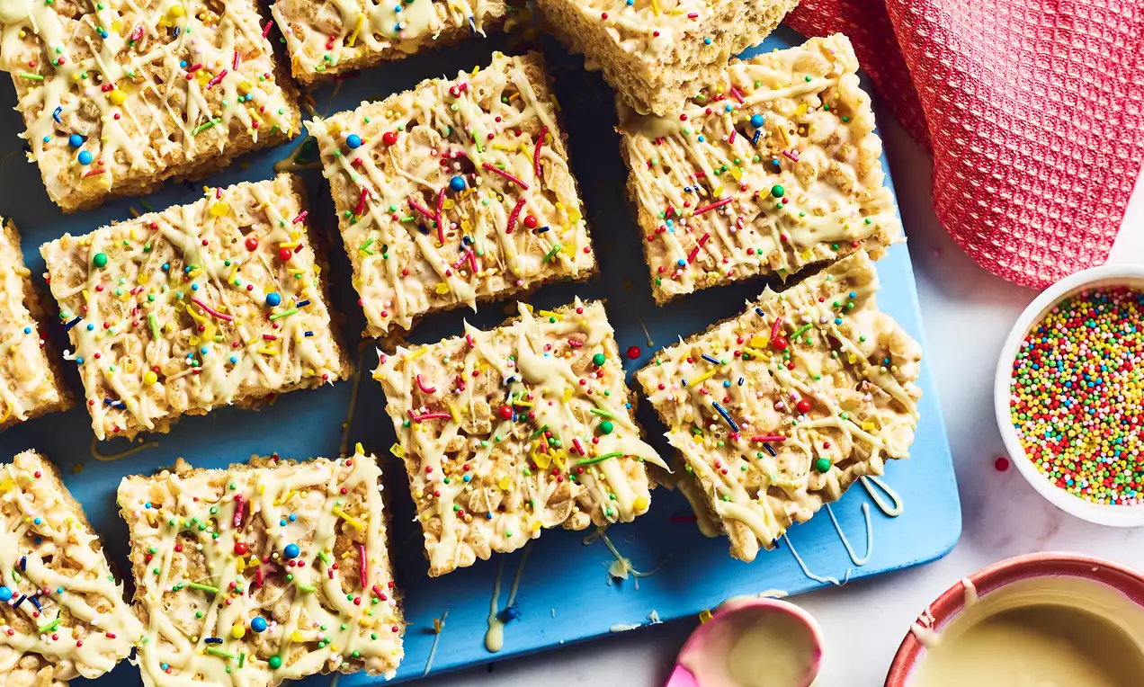 Golden square-shaped Mallow Crispy Treats topped with drizzles of white icing and colourful sprinkles on a blue board
