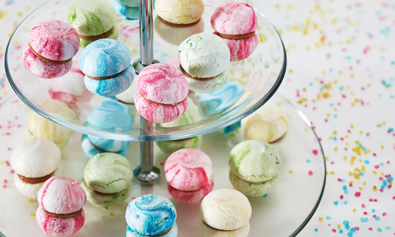 Bunte Meringues in pastellfarbenem Marmormuster, rund und luftig, auf einer gläsernen Etagere angeordnet, umgeben von farbigem Streudekor