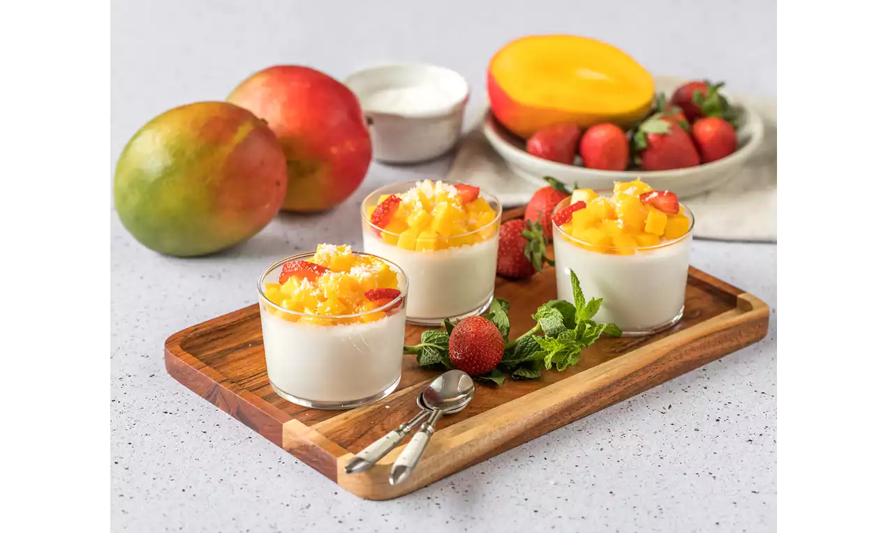 Vasitos de crema blanca suave coronados con cubos de mango amarillo brillante, fresas cortadas y coco rallado sobre una tabla de madera