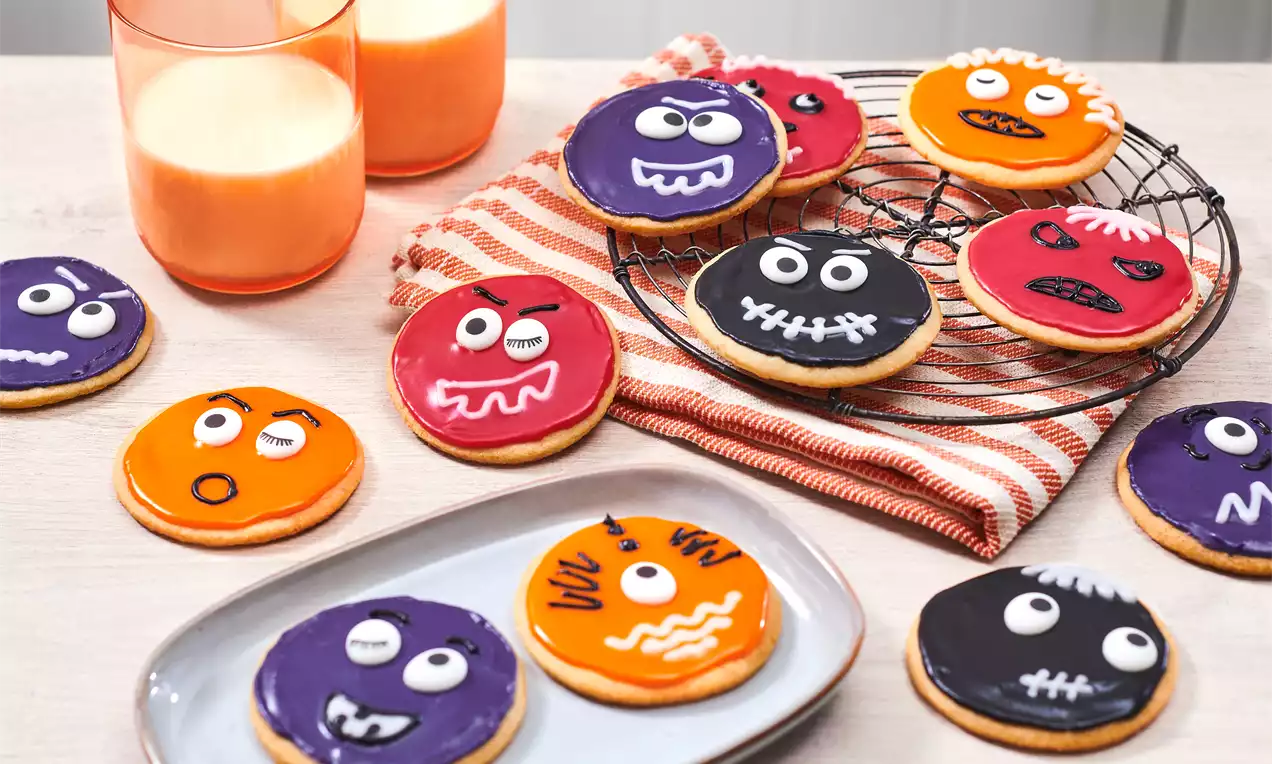 Biscuits ronds aux glaçages colorés orange, violet, rouge et noir, décorés de visages effrayants sur une grille et une assiette