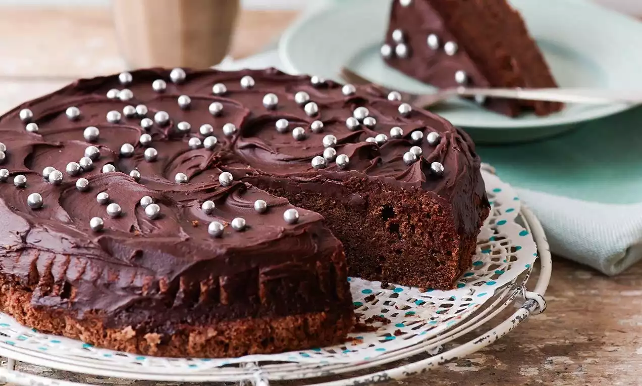 Mørkebrun sjokoladekake med glinsende sjokoladetrekk, pyntet med små sølvkuler, servert på dekorativt fat med en oppskåret bit i bakgrunnen