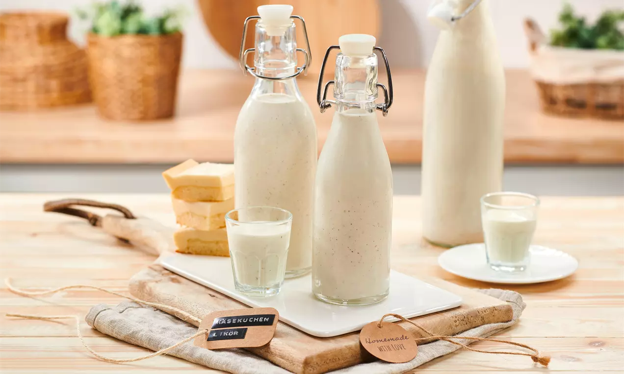 Käsekuchen-Likör in schlichten Glasflaschen auf weißer Platte mit Gläsern, gelben Kuchenstücken und Dekoration aus Holz und Stoff drapiert