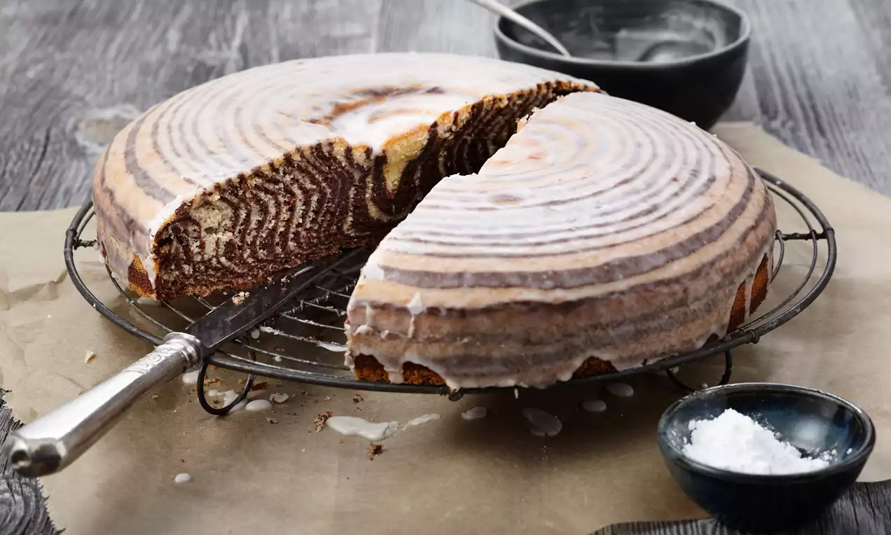 Runder Zebrakuchen mit spiralförmigem Muster und weißem Zuckerguss, auf Metallgitter angerichtet, umgeben von Schalen und Messer