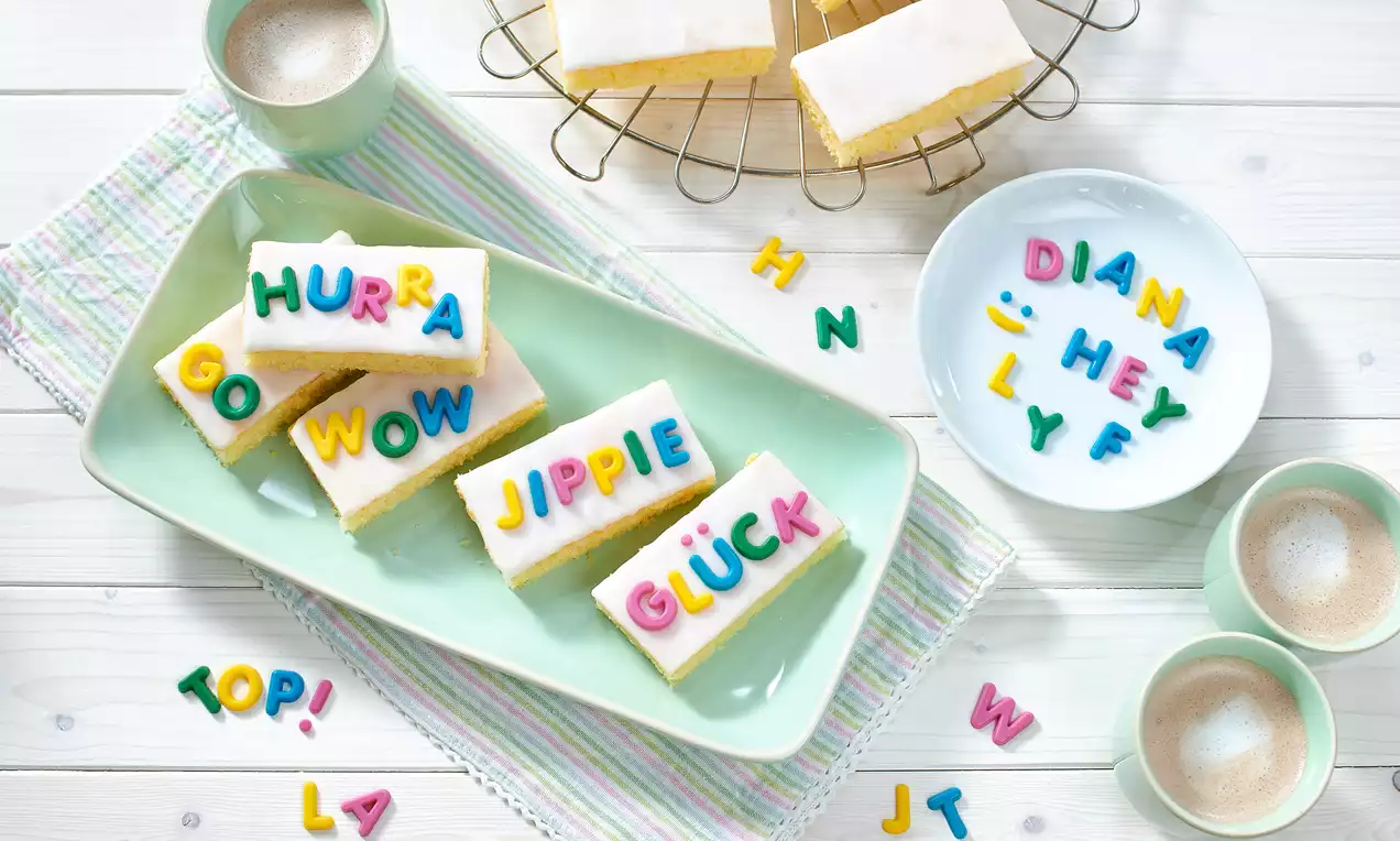 ABC-Kuchen mit heller Glasur, bunte Buchstaben aus Zuckerguss, auf grüner Platte, umgeben von bunten Buchstaben und Geschirr auf Holzuntergrund