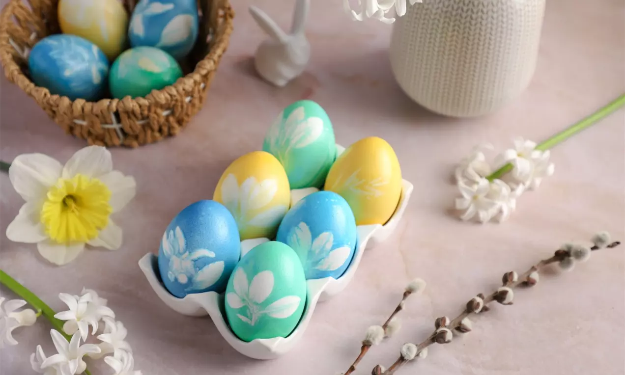 Oslikana uskršnja jaja pastelno plavih, zelenih i žutih tonova sa belim cvetnim motivima u keramičkom poslužavniku, okružena svežim cvetovima
