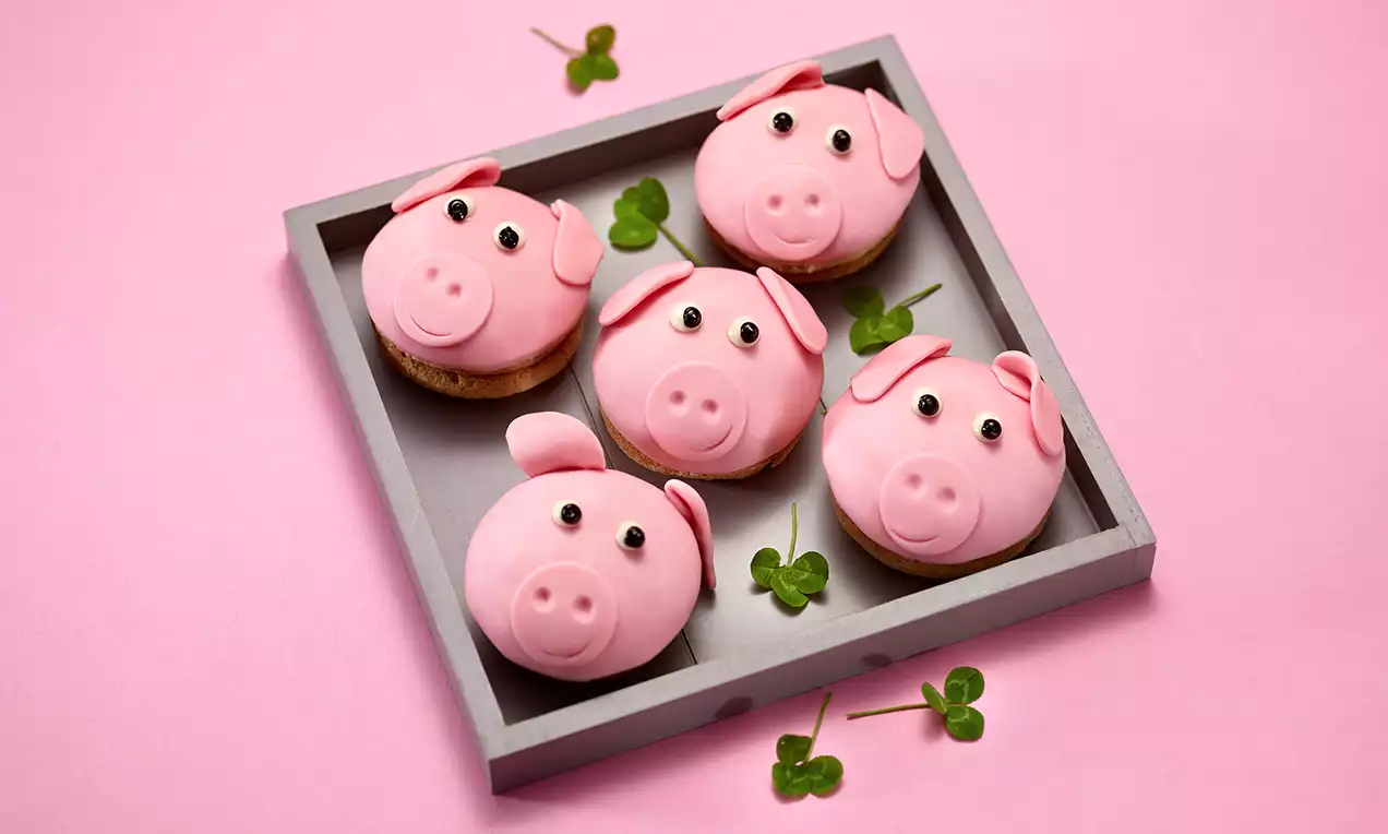 Rosa Schweinchen-Cupcakes mit glatter Fondant-Oberfläche, schwarzen Augen und Ohren auf grauem Tablett, verziert mit grünen Kleeblättern