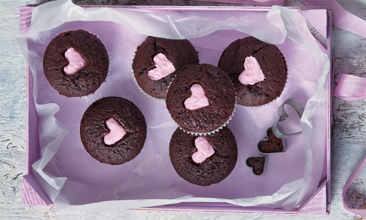 Čokoládové muffiny s matnou hnědou krustou, zdobené růžovými cukrovými srdíčky, na fialovém podnosu s jemným bílým pergamenem