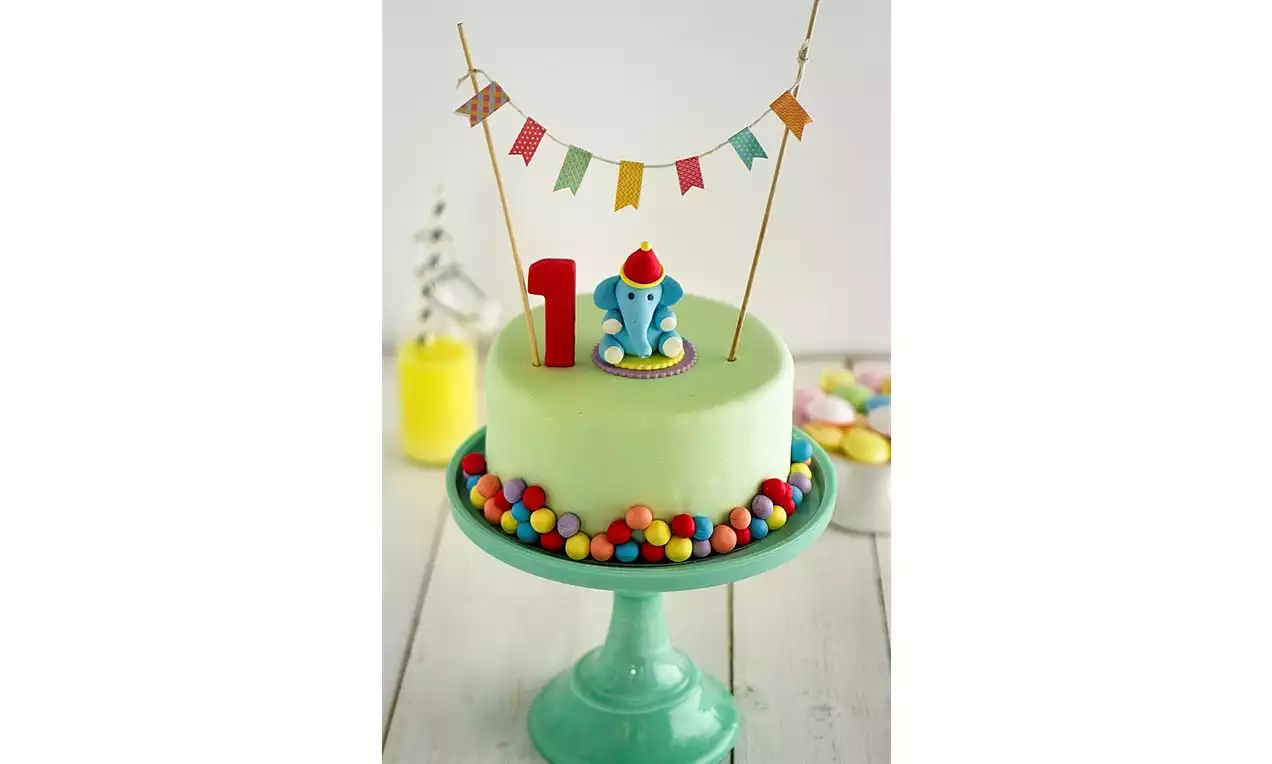 Rund, lysegrønn kake på grønt kakefat, dekorert med fargerike kuler, elefantfigur, rød ett-tall og flaggrekke i trekanter