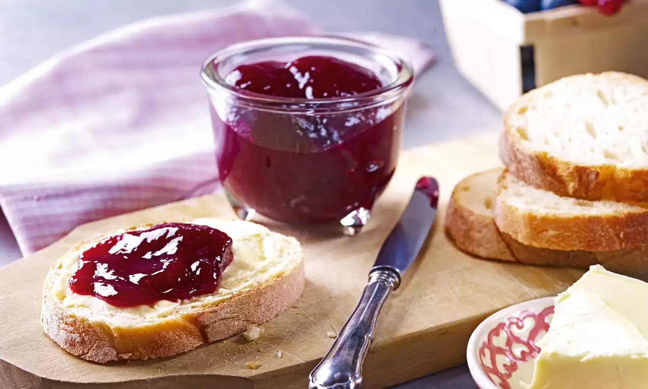 Samtig rote Heidelbeer-Johannisbeer-Konfitüre im Glas, daneben bestrichenes Brot, Butter, Messer und Beeren in hellem, natürlichem Licht