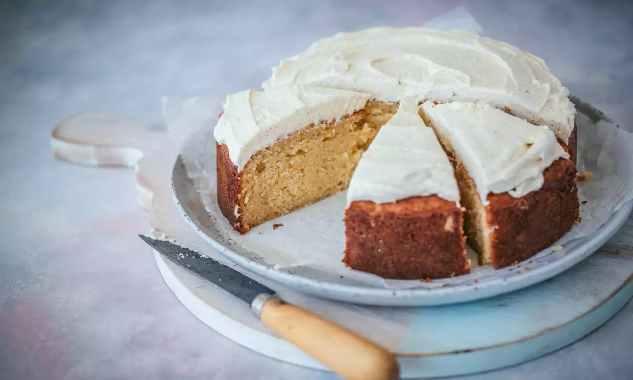 Tort cu cremă de unt vanilată albă, cu blat auriu pufos, feliat pe un platou rotund alb, lângă un cuțit cu mâner din lemn