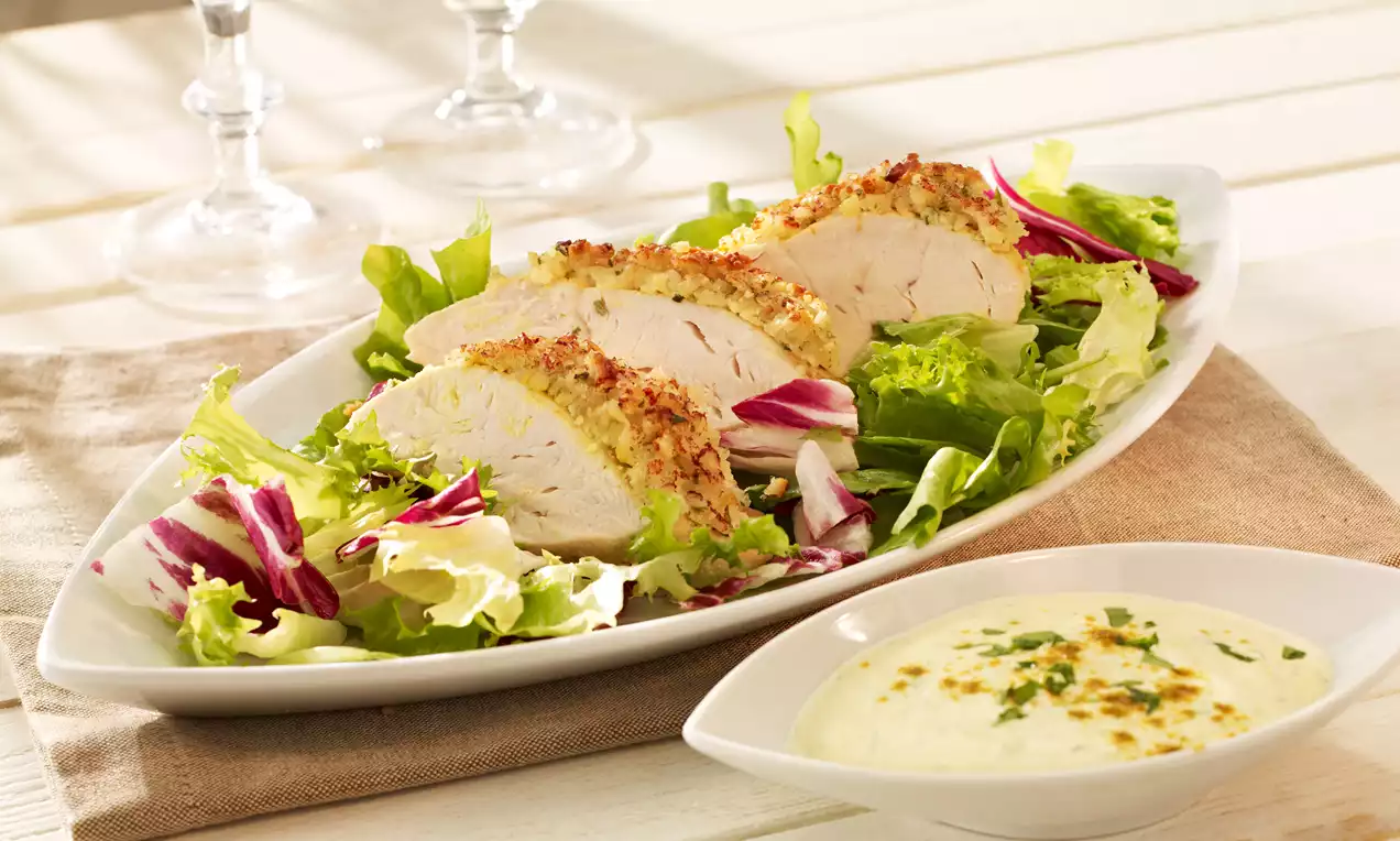 Saftige Hähnchenbrust mit goldbrauner Mandelkruste auf gemischtem Salat, serviert auf weißer Platte neben einer cremigen hellen Soße