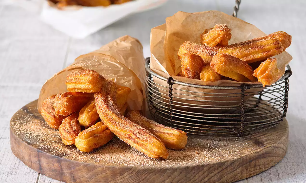 Zlatavé churros s křupavou skořicovo-cukrovou krustou uspořádané na dřevěné desce, část v drátěném košíku a část ve hnědém papíru