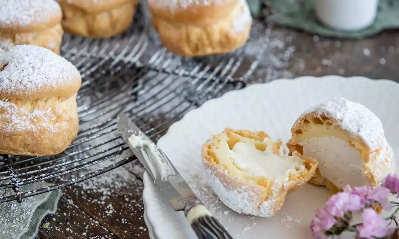 Picture - Golden vanilla profiteroles dusted with fine white icing sugar, some on a cooling rack and others on a white plate with pink flowers