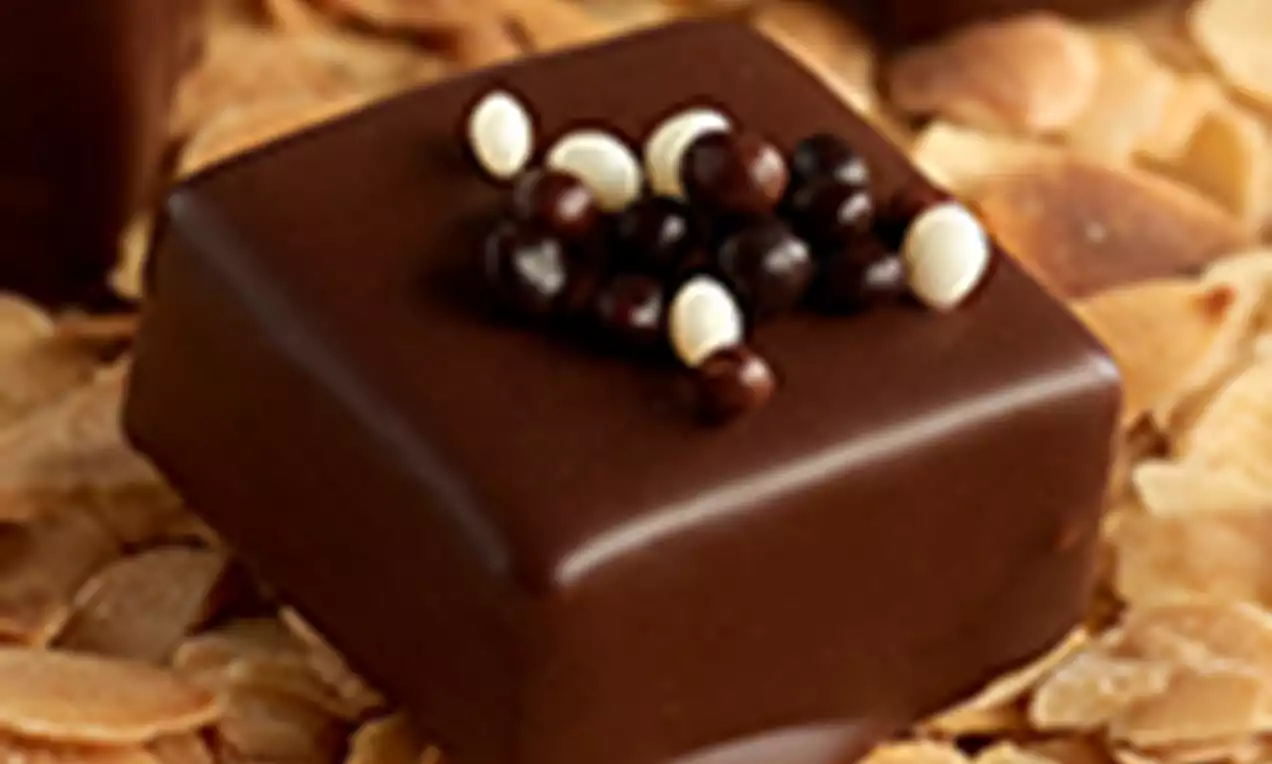 Cuadrados de mazapán cubiertos de chocolate brillante con pequeñas perlas blancas y negras sobre una cama de láminas de almendra dorada